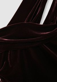 Burgundy bársonydressz, lágy textúrájú, áramló anyaggal és redőzött nyakkivágással, amely kiemeli a ruhadarab elegáns kialakítását.