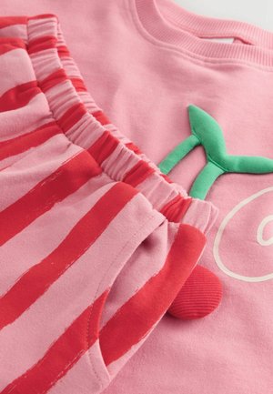 Rosa Sweatshirt mit einem grünen Applikationsdetail, kombiniert mit rosa Shorts mit roten Streifen. Weicher Stoff, elastischer Bund und runder Akzent.