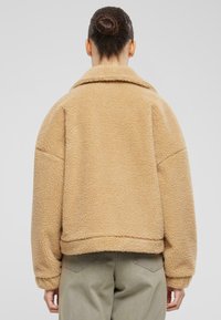 Giacca in pile beige oversize con superficie texturizzata, colletto arrotondato e polsini a coste, abbinata a pantaloni di color chiaro.