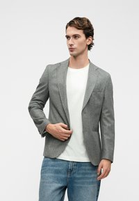 Šedý texturovaný blazer s přiléhavým střihem, jednopřezkovým zapínáním a klopami, oblečený přes bílé tričko a pohodlné modré džíny.