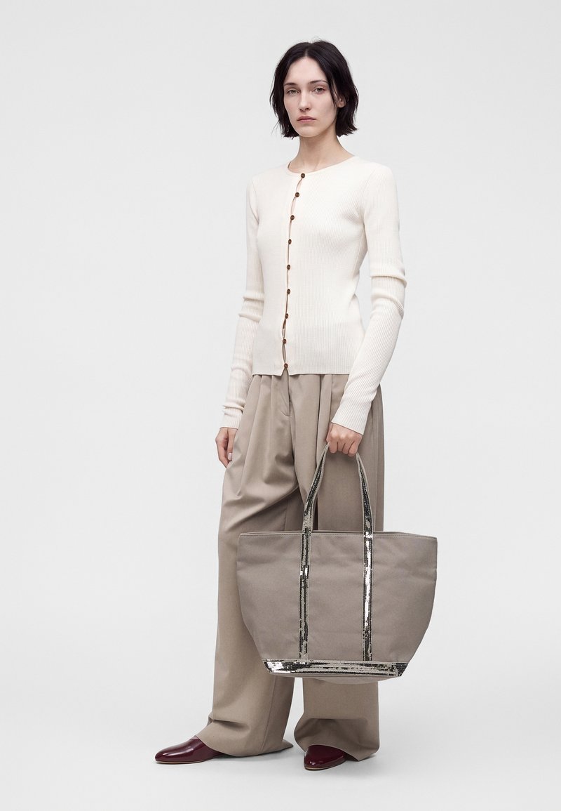 Femme portant un cardigan blanc boutonné et un pantalon large beige tenant un grand sac cabas beige avec des poignées et des accents argentés brillants.