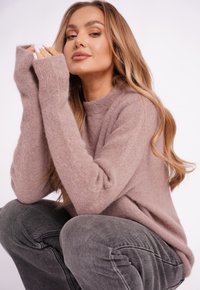 Jasnoróżowy, puszysty sweter z długimi rękawami i zaokrąglonym dekoltem; w połączeniu z szarymi dżinsami. Modelka kuca, prezentując oba ubrania.