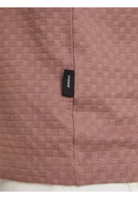 Zbliżenie teksturowanej brązowej tkaniny z małym kwadratowym wzorem oraz czarną metką z napisem "CHASIN'" wszytą w pobliżu szwu nad białą tkaniną.