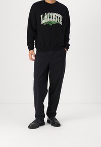Felpa nera con logo verde "LACOSTE", abbinata a pantaloni neri larghi e scarpe nere con suole texturate.