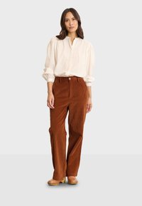Pantaloni in corda marrone a coste lunghe con vita alta, abbinati a una camicia bianca con bottoni e zoccoli marroni. Tessuto liscio e strutturato.