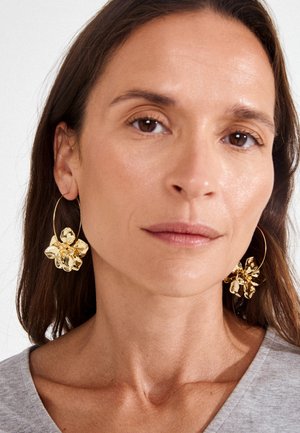 Žena s dugom smeđom kosom koja nosi velike zlatne naušnice u obliku cvijeta i sivu majicu, gleda ravno sa neutralnim izrazom.