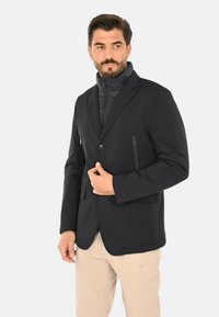 Blazer nero dal design elegante, con chiusura a bottoni, due tasche frontali con zip e un interno a collo alto a più strati.