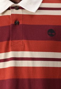 Rdeče in kremnoebeljn skupini polo majica z belim ovratnikom. Ima dva rjava gumba in vezeno drevesno logo na levi strani.