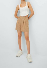 Vit kortärmad tanktopp med fyrkantig halsringning, tillsammans med beige veckade shorts och vita sneakers, mot en ljusgrå bakgrund.