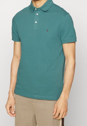 Mężczyzna noszący turkusową koszulkę polo z krótkim rękawem, trzema białymi guzikami i małym logo Tommy Hilfiger, w połączeniu z beżowymi wzorzystymi spodniami.