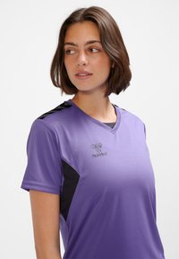 Lila sporttröja med korta ärmar, V-ringning, mörka sidopaneler och en logotyp på bröstet. Tyget verkar vara slätt och andningsbart.