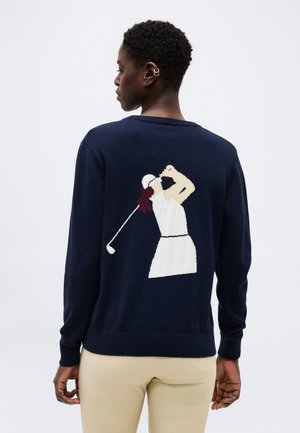 Persoon met kort haar draagt een donkerblauwe trui met een golfer in volle zwaai op de rug, gecombineerd met lichtbeige broek.