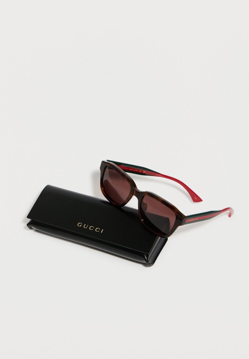Lunettes de soleil œil de chat en écaille de tortue avec des branches rouges et vertes reposant sur un étui Gucci noir avec logo doré sur fond blanc.