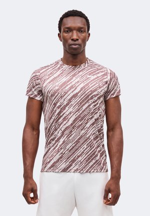 Homme de face portant un T-shirt à manches courtes rayé abstrait marron et blanc et un short blanc sur un fond uni.