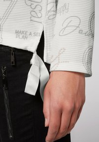 Camisa blanca de manga larga de canalé con patrones de texto impresos. El dobladillo cuenta con un detalle de cinta atada, combinada con pantalones negros con cremallera.