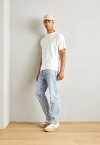 Afslappet outfit bestående af en cremefarvet t-shirt, lyseblå jeans og beige sneakers med teksturerede detaljer. En beige kasket fuldender looket.