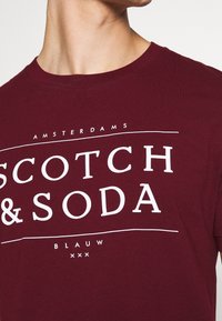Mørkerød bomulds t-shirt med hvid tekst "SCOTCH & SODA", med subtile accentlinjer og sætningerne "AMSTERDAMS" og "BLAUW." Klassisk rund hals.
