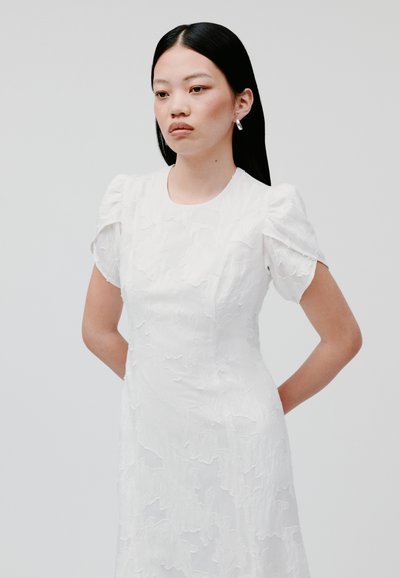 Abito bianco con motivo floreale testurizzato, maniche a palloncino corte e scollatura rotonda. La modella posa con le mani incrociate dietro la schiena.