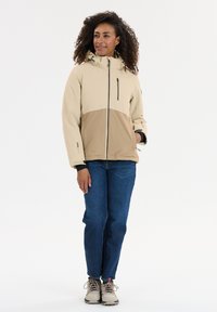 Beige en bruine kleurblokjas met een capuchon, voorzien van een rits aan de voorkant en een ritsborstzak, in combinatie met blauwe jeans en lichtgekleurde schoenen.