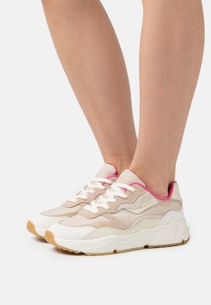 Füße, die beige und weiße Sneakers mit pinkem Besatz und weißen Schnürsenkeln auf hellem Hintergrund tragen, zeigen Seiten- und Vorderansichten.