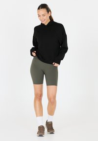 Zwarte hooded sweatshirt, relaxte pasvorm, gedragen met olijfgroene fietsshorts. Bruine sneakers en witte crewkousen maken de outfit compleet.