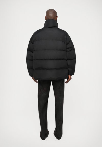Zwarte pufferjack met horizontale gestikte naden, hoge kraag en een losse pasvorm. Het materiaal lijkt glad en glanzend en is gecombineerd met donker gestreepte broek.