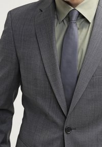 Grauer Blazer mit strukturierter Oberfläche, kombiniert mit einem hellgrünen Hemd und einer dunkelgrauen Krawatte. Ein-Knopf-Verschluss am Blazer.