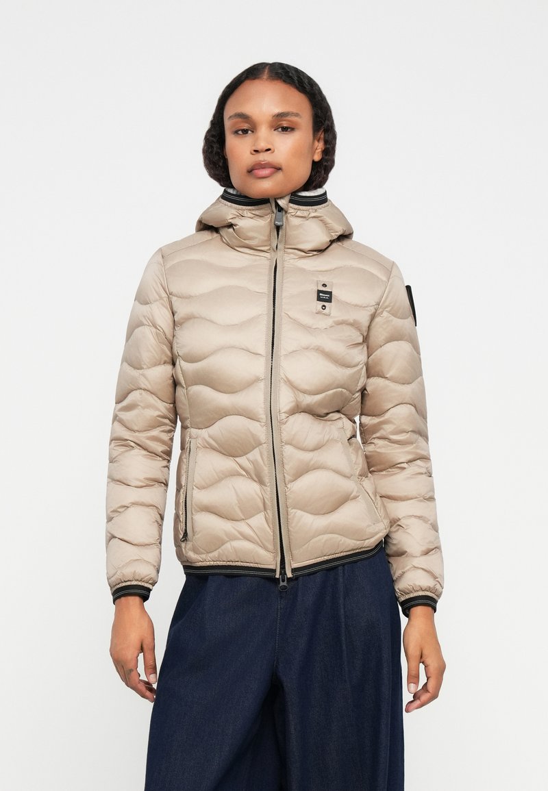 Jachetă puffer bej cu glugă, design matlasat, fermoar frontal, două buzunare laterale și manșete canal. Prezintă un eticheta cu logo pe piept.