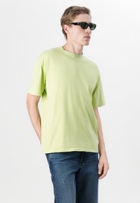 Light green cotton t-shirt with short sleeves, relaxed fit, crew neck, and subtle branding on the front. Worn with dark denim jeans.