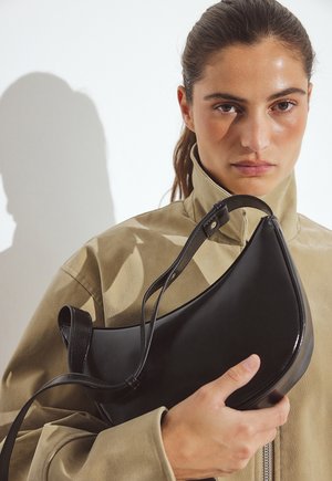 Zwarte leren tas met een gebogen vorm, met een lange schouderband en een gladde textuur, vastgehouden door een persoon in een beige jas.