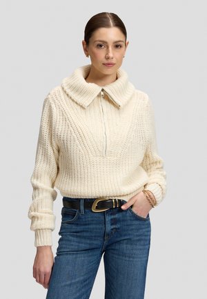 Kvinde iført en cremefarvet chunky strikket sweater med bred krave og lynlås, blå jeans, sort bælte med guldfarvet spænde og guldbelagte armbånd.