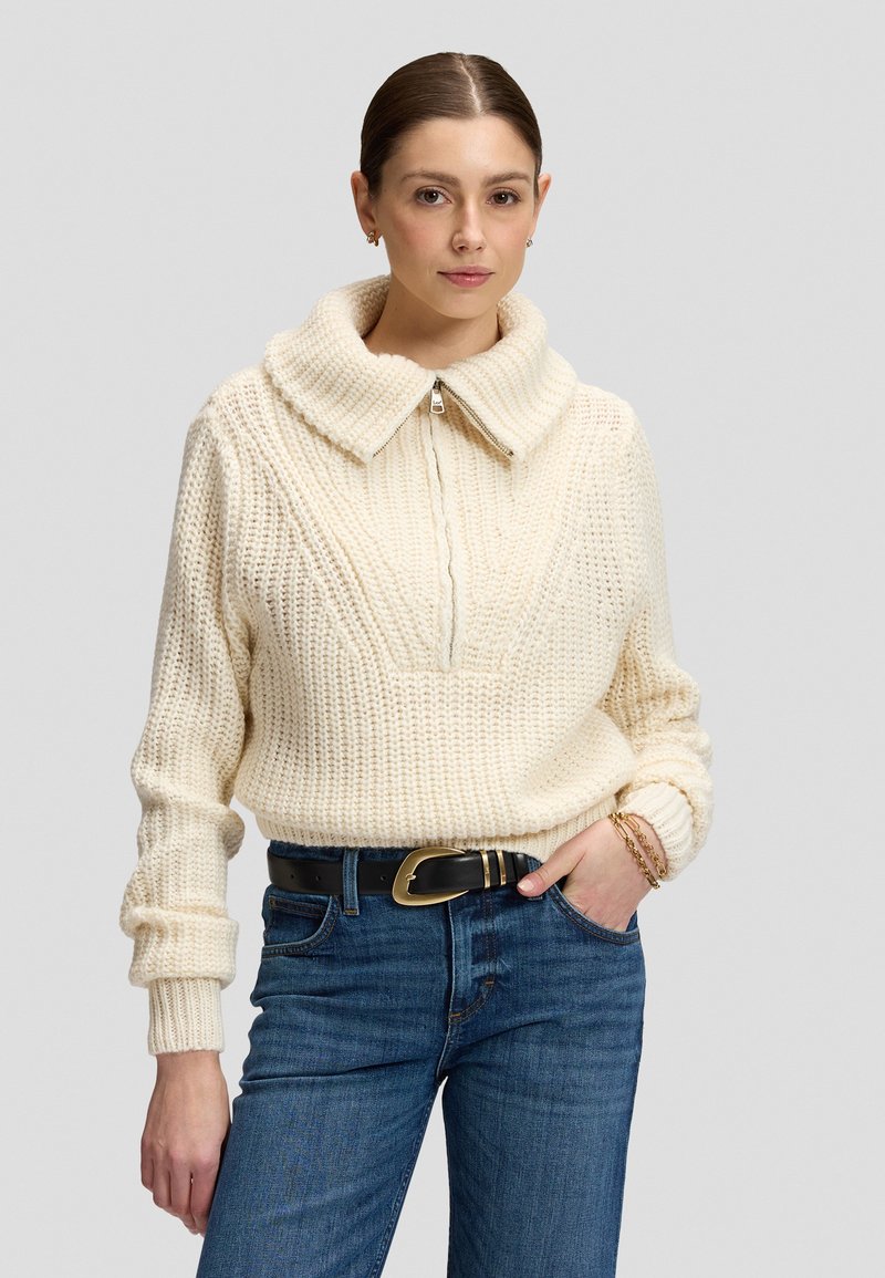 Femme portant un pull en grosse maille couleur crème avec un large col et une fermeture éclair, un jean bleu, une ceinture noire avec une boucle dorée et des bracelets dorés.