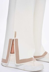 Witte gebreide broek met beige accenten, voorzien van een zijsplit en een merklabel, gecombineerd met een witte sneaker met subtiele slijtageplekken.