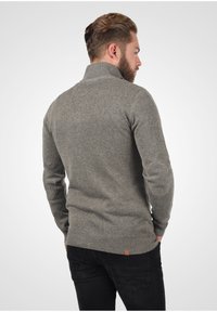 Maglione grigio lavorato a maglia con colletto alto, polsini e orlo a coste. Il retro presenta una superficie liscia e un marchio minimo vicino al bordo inferiore.