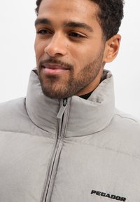 Graue Steppjacke aus weichem Stoff mit hohem Kragen, vorne mit Reißverschluss und einem kleinen schwarzen Logo auf der Brust.