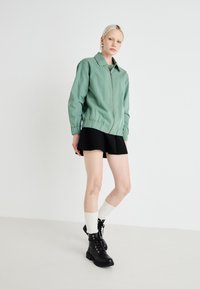 Giacca verde con zip, dal collo alla coreana e polsini elasticizzati, abbinata a una gonna nera svasata, calze bianche fino al ginocchio e stivaletti neri con lacci.