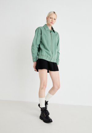 Green, zip-up jacket with a collared neckline and elastic cuffs, paired with a black flared skirt, knee-high white socks, and black lace-up boots.
