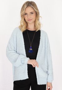 Cardigan lavorato a maglia di un azzurro chiaro, soffice e con una vestibilità ampia, spalle scese, polsini a coste e apertura frontale. Indossato sopra un top nero, abbinato a una collana con nappina blu.