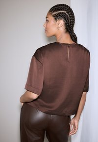 Blusa marrone in raso con maniche corte arrotolate, con dettaglio a goccia sulla schiena e vestibilità morbida, abbinata a pantaloni in pelle marrone coordinati.