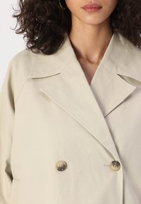 Beige double-breasted coat with large lapels, featuring two round, tan buttons and a smooth, matte texture.