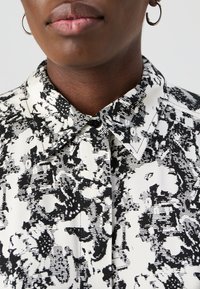 Svart-hvitt mønstrete bluse med klassisk krage, knappelukking, og en jevn tekstur, med abstrakte design.