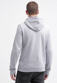 Mężczyzna widziany z tyłu, ubrany w jasnoszary sweter z kapturem i czarne dżinsy, na jednolitym białym tle.