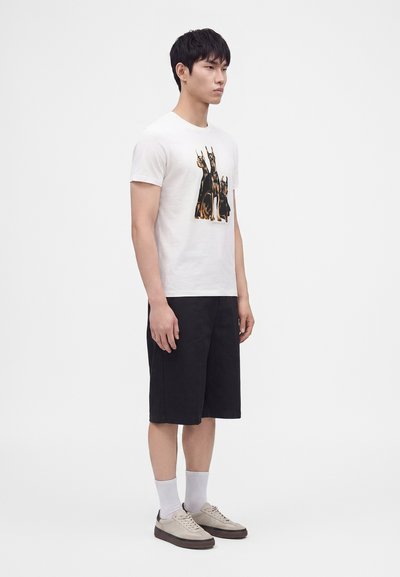 Άνδρας που φοράει λευκό T-shirt με σχέδιο τριών σκύλων Doberman, μαύρα σορτς μέχρι το γόνατο, λευκές κάλτσες και μπεζ αθλητικά παπούτσια, στέκεται πλαγίως.