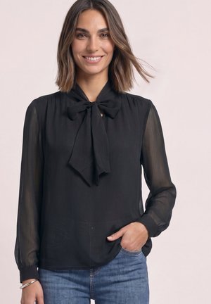Femme souriante portant un chemisier noir avec un nœud au col et des manches longues transparentes, une main dans la poche d’un jean bleu.