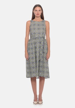 Donna che indossa un vestito senza maniche lungo fino al ginocchio con un motivo geometrico verde, blu e nero, in piedi di fronte con le braccia rilassate ai lati.