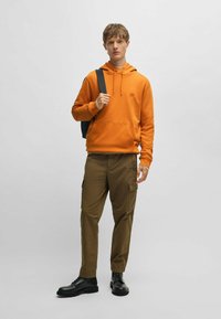 Oranje hoodie met een buidelzak aan de voorkant, gecombineerd met khaki cargobroeken met zijzakken en zwarte veterschoenen.