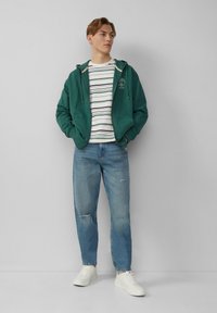 Groene katoen hoodie met rits, T-shirt met witte en marineblauwe strepen, lichtblauwe versleten jeans en witte sneakers. Eenvoudig ontwerp, casual uitstraling.