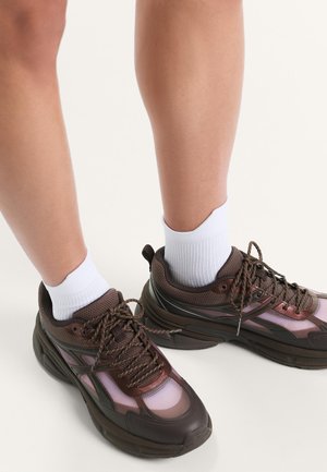 Benen met witte enkelsokken en stevige bruine sportschoenen met zwarte zolen op een effen lichte achtergrond.