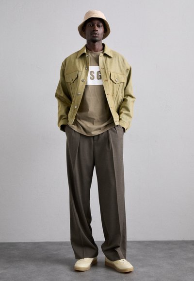 Khaki cord jacket with two chest pockets, layered over a taupe t-shirt featuring white logo. Loose brown trousers and cream sneakers complete the look.