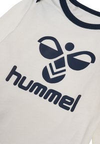 Vit t-shirt med en marinblå logotyp som har abstrakta former och varumärkesnamnet "hummel" tydligt angivet längst ner.